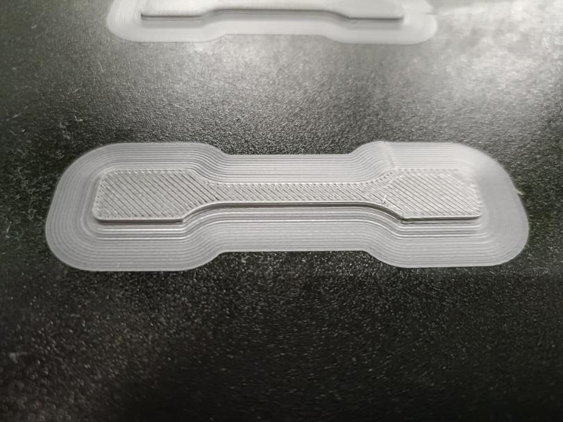 Original photograph of a white dog-bone-shaped tensile-test specimen on a printer bed
